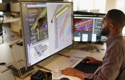 A man sitting at a desk looking at a PC screen showing various building schematics.